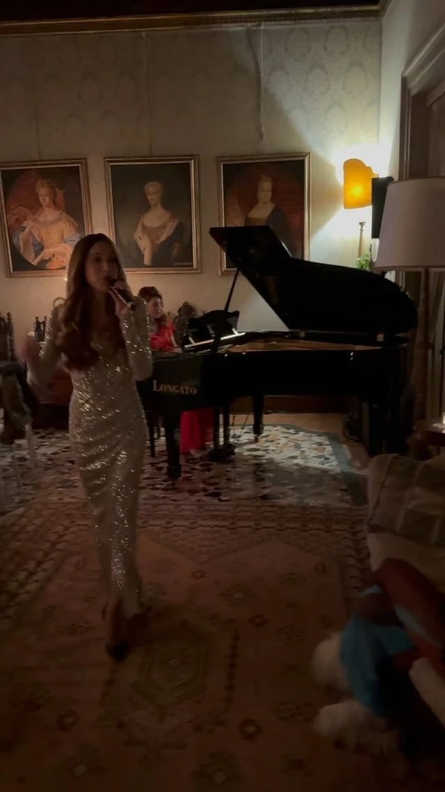 A singer beside a grand piano in a salon hung with portraits