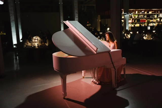 A white grand piano lit in pink in a dark hall, guests dining in the background