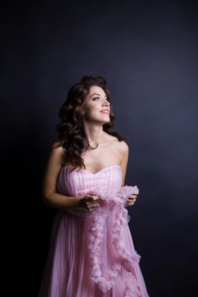 Giulia Vazzoler in a strapless blush-pink tulle gown, smiling and looking up, against a dark backdrop