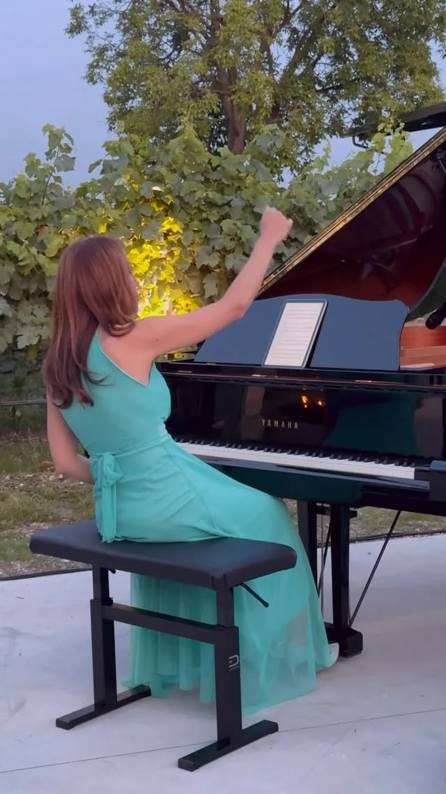 A pianist in green playing a grand piano outdoors under trees