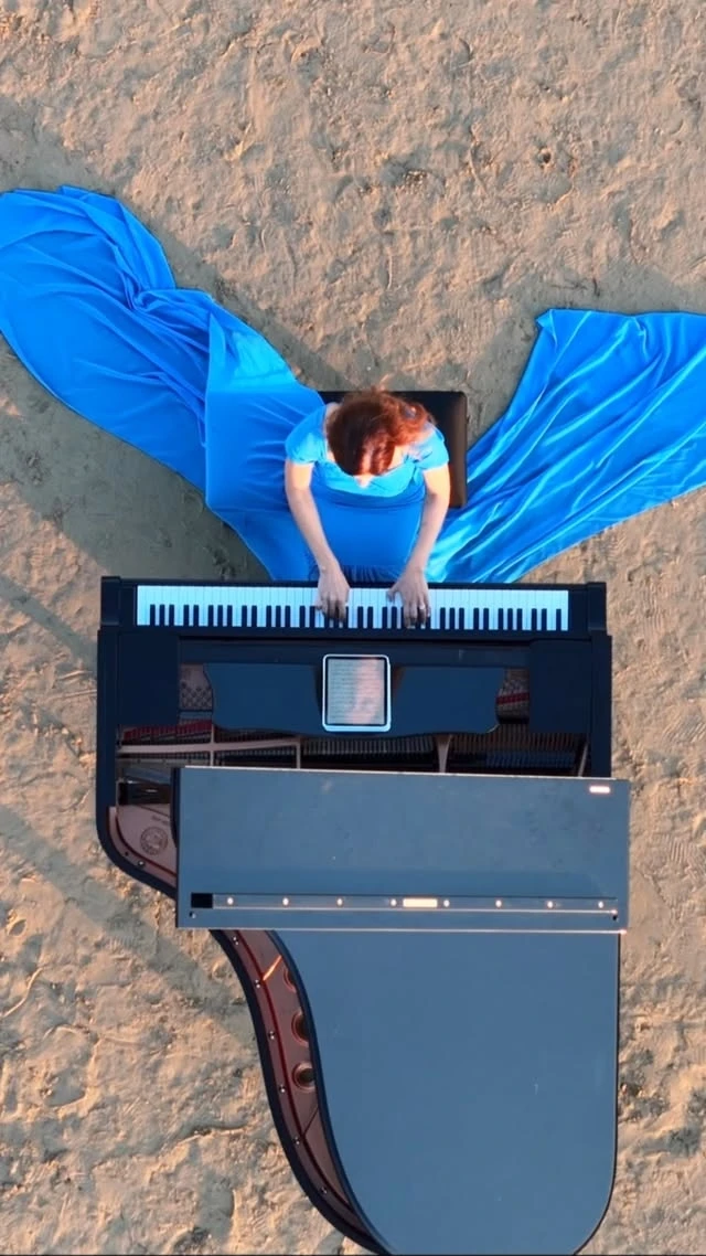 A pianist in a flowing blue dress at a black grand piano, seen from above