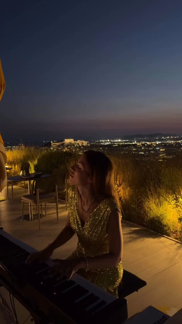 A pianist at a keyboard on a terrace at dusk, the lights of a city below